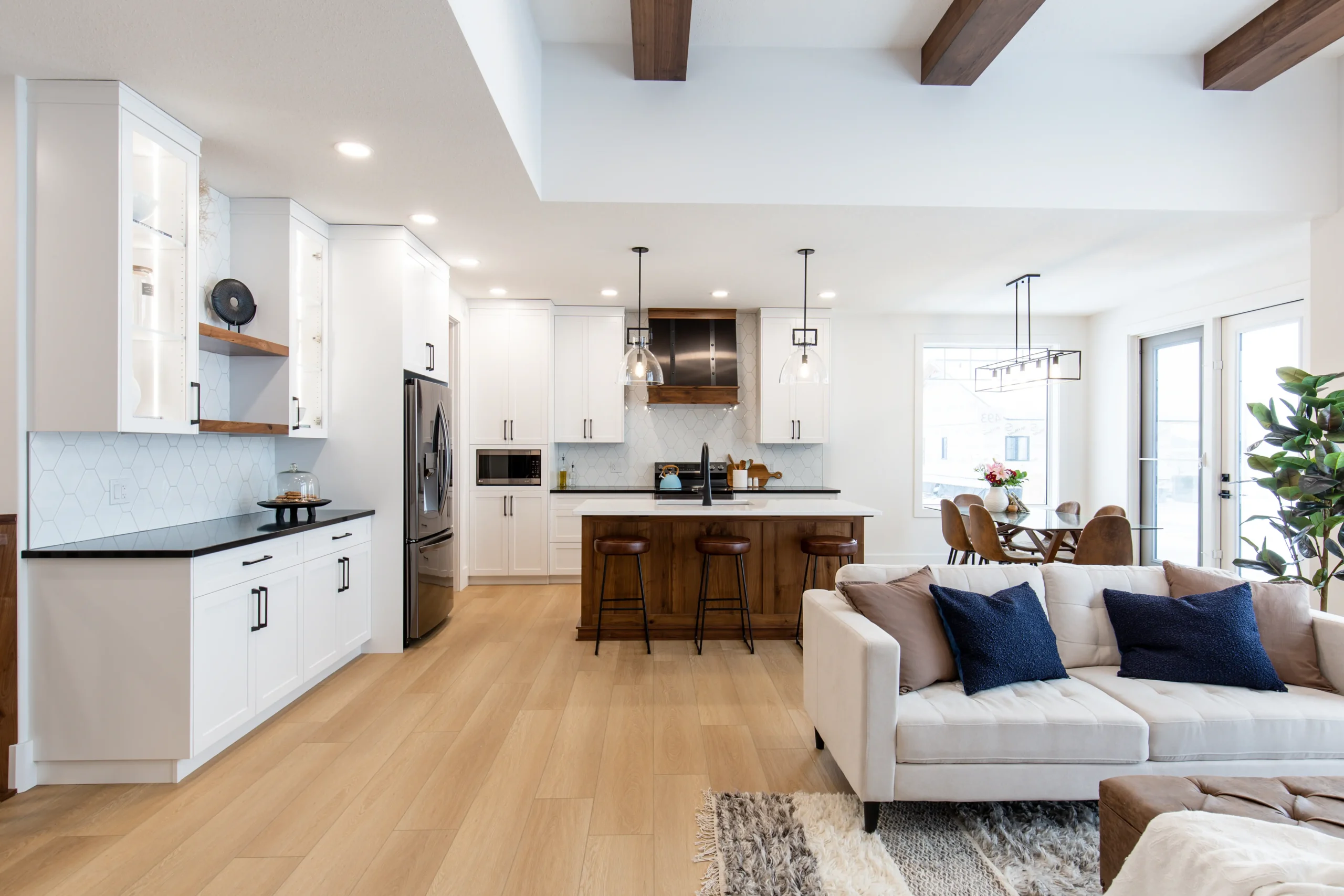 Open-concept interior of a ready-to-move (RTM) home featuring a modern kitchen with white cabinetry, wood island, stainless steel appliances, pendant lighting, and dining area connected to a bright living room with exposed ceiling beams.