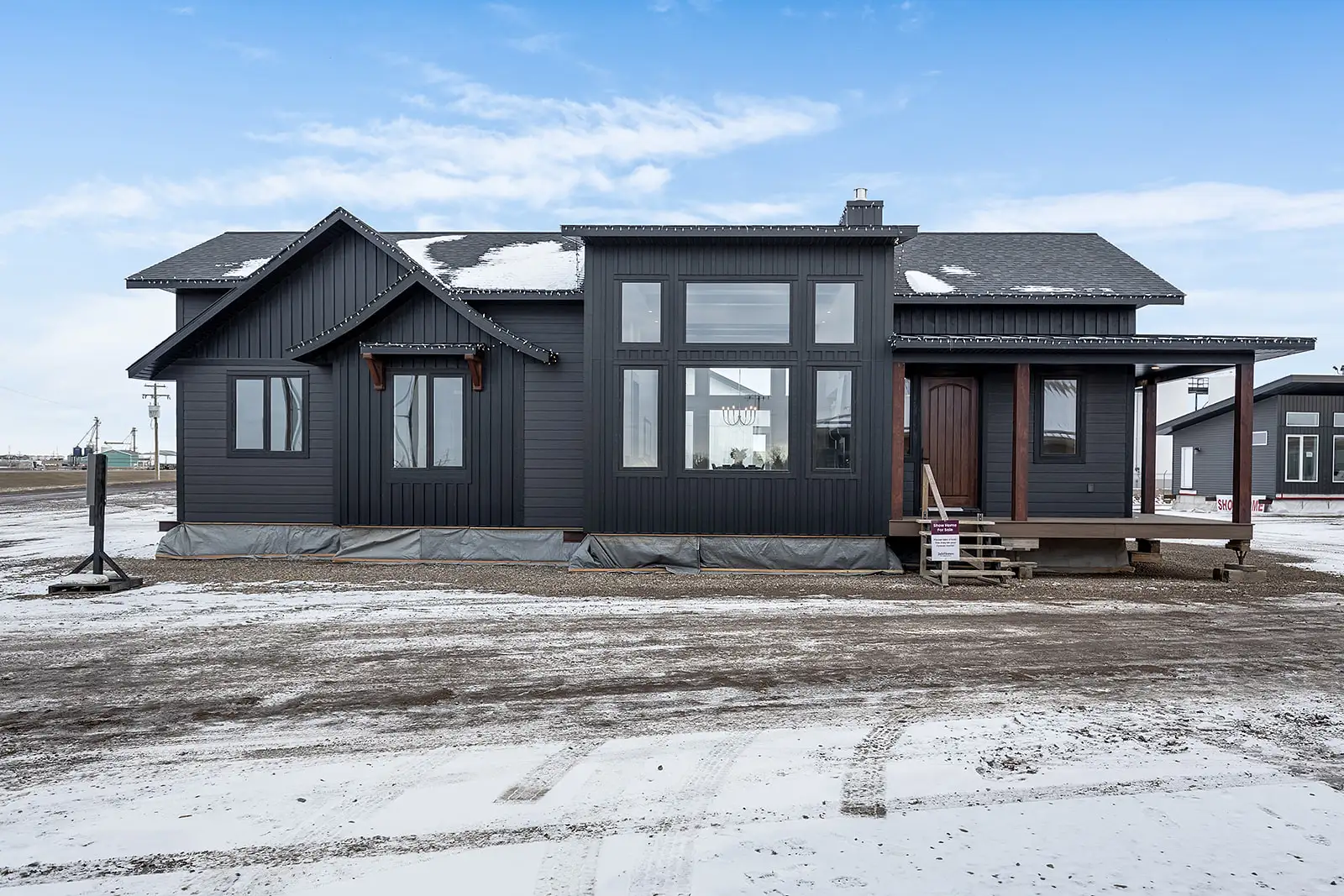 a ready-to-move (RTM) home with dark gray siding and large windows, set on a snowy lot. The home features a high-pitched gable roof and a covered front porch with wooden posts, adding warmth to the sleek exterior. The snow-dusted roof and the surrounding ground indicate a cold, wintry setting. The large windows provide a view of the well-lit interior, including a chandelier. The home is slightly elevated, awaiting transport to its final destination. Other RTM homes are visible in the background, set against a partly cloudy sky.