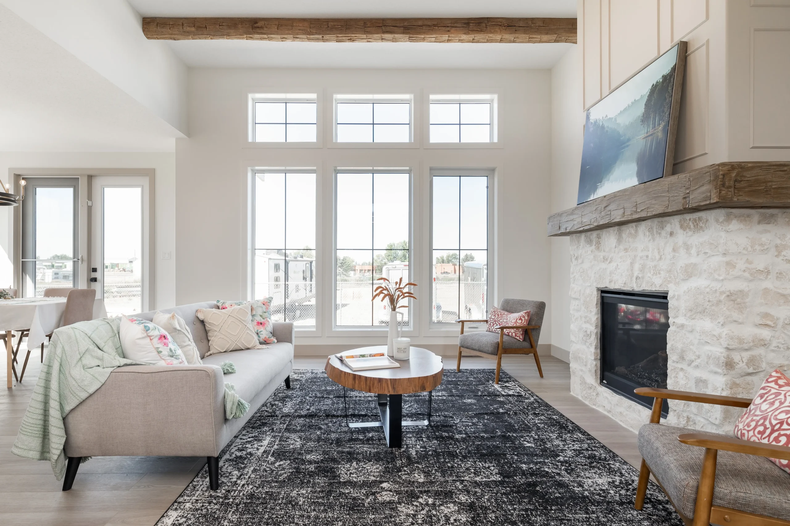 A bright and modern living room featuring a large stone fireplace with a wooden mantel, topped by a flat-screen TV. The space is decorated with neutral tones and floral accent pillows on a light gray sofa. The seating area also includes a pair of mid-century modern armchairs and a wooden coffee table set on a textured black and white area rug. The room is filled with natural light from large, grid-patterned windows, and an exposed wooden beam adds a rustic touch to the otherwise contemporary design. The open-concept layout extends to a dining area in the background, creating a welcoming and airy atmosphere.