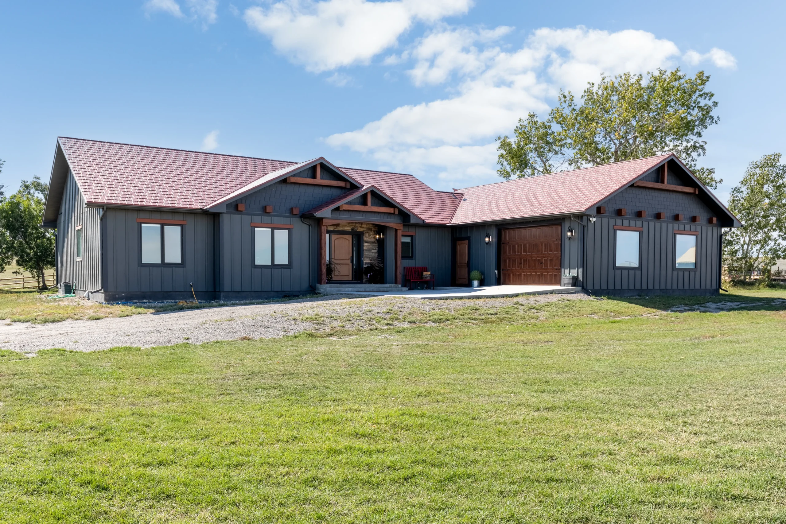 A modern single-story home with dark gray siding and a red metal roof, featuring a large, welcoming front porch with wooden accents. The house has a double garage with wooden doors and several large windows that allow natural light inside. The front yard is an expansive grassy area with a gravel driveway leading to the garage and front entrance. The architectural design includes gabled roofs and timber beams, giving the home a rustic yet contemporary appearance. Trees surround the property, set against a bright blue sky with scattered clouds, enhancing the peaceful, rural setting.