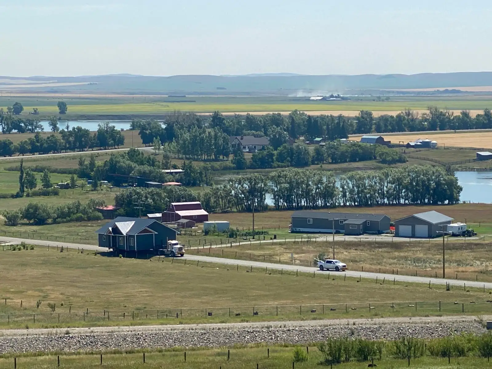 A scenic rural landscape in Alberta showing a ready-to-move (RTM) home being transported by a truck on a country road. The surrounding area features expansive fields, trees, and distant farmlands under a clear blue sky. Several homes, barns, and outbuildings are visible, along with a small body of water reflecting the peaceful rural setting. A white pickup truck follows behind the house transport, and the rolling hills in the distance add depth to the serene and picturesque countryside view.