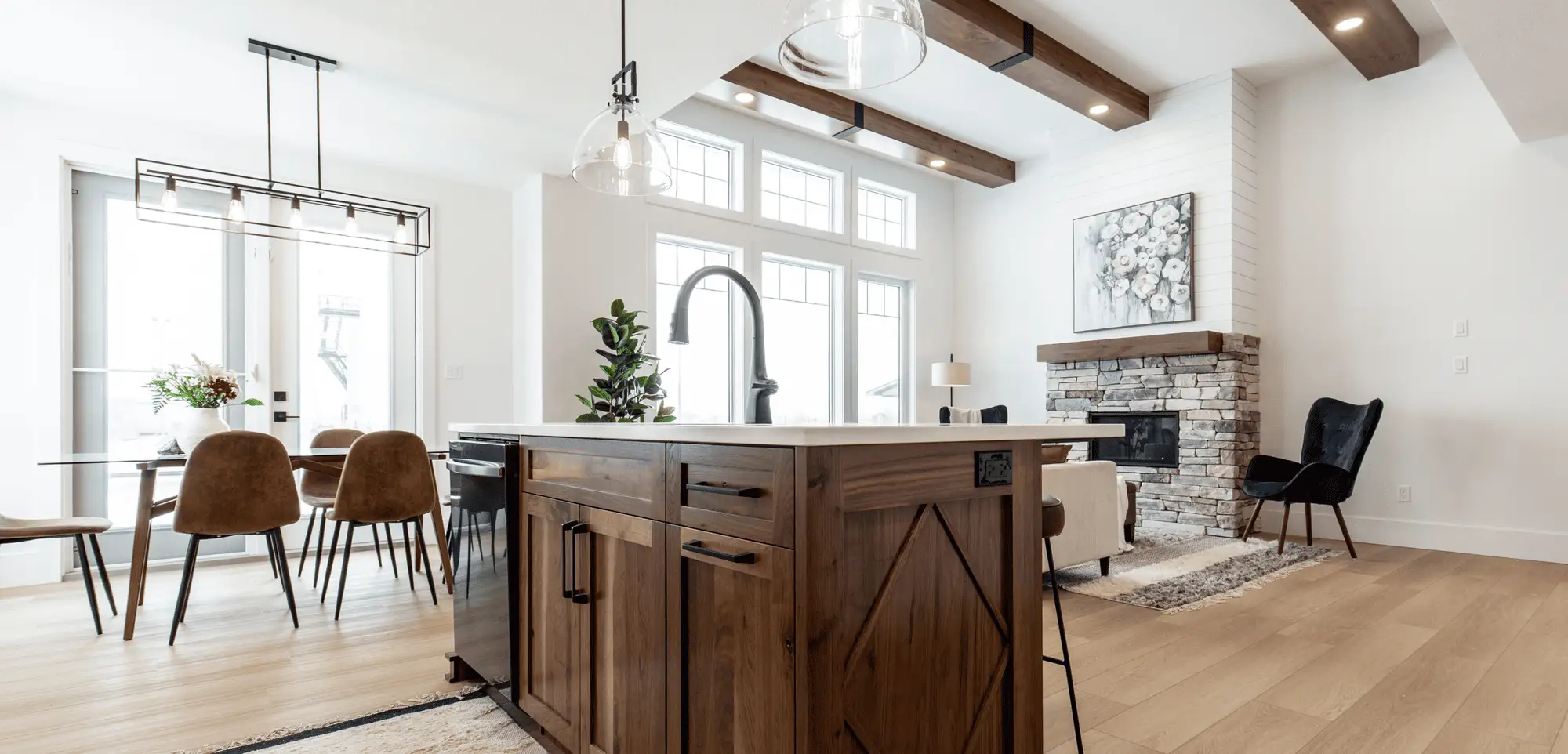 Open-concept RTM great room featuring a wood kitchen island with pull-down faucet, grid windows under exposed beams, stone-and-shiplap fireplace with wood mantel, glass pendants, and wide plank floors.