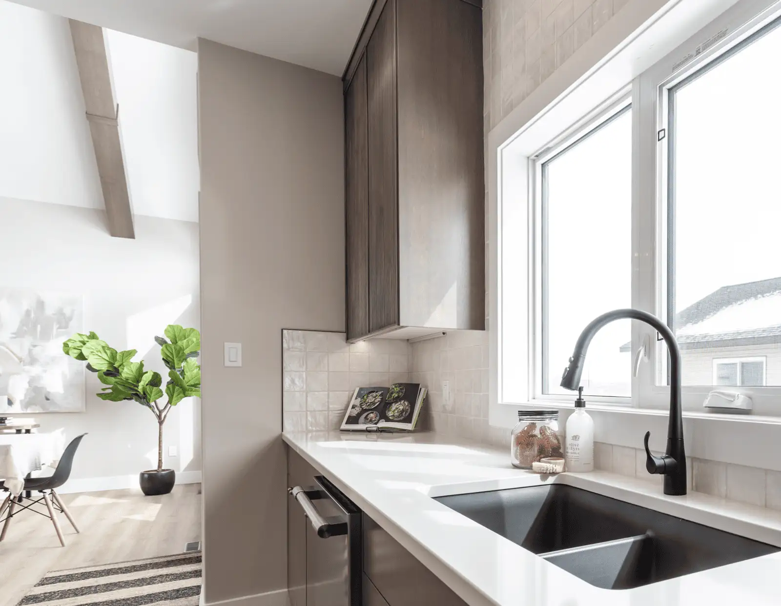 Kitchen close-up with black pull-down faucet and undermount sink in white quartz countertop, square tile backsplash, tall wood cabinets, and a glimpse of the dining area with a fiddle-leaf fig.