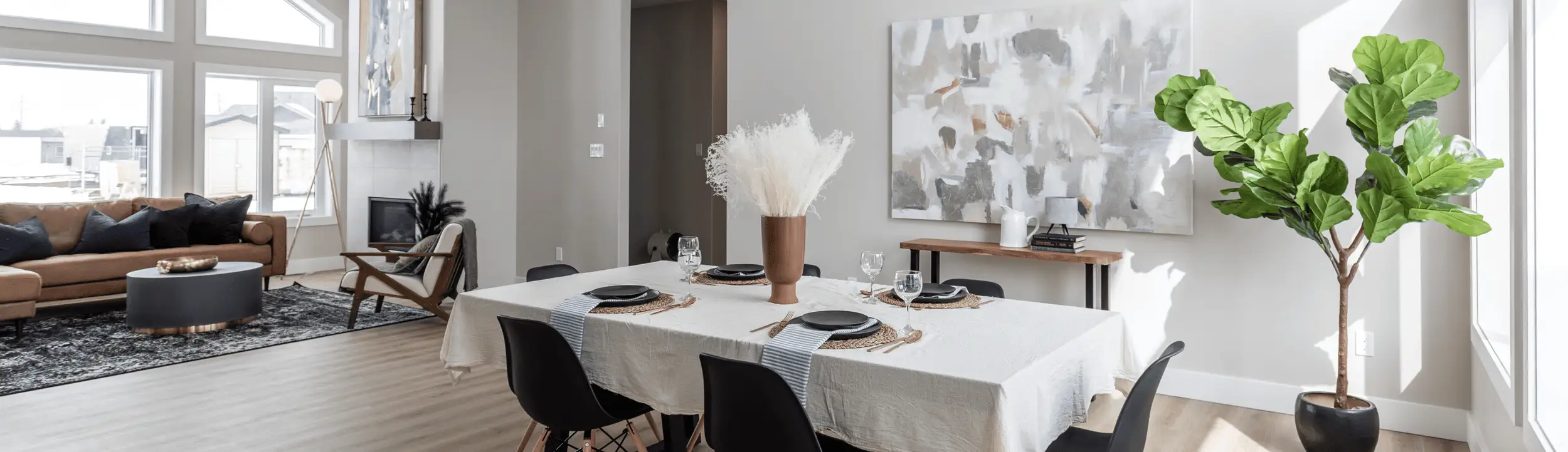 Sunlit dining area with a table set in black dinnerware and a pampas-grass centerpiece, large abstract artwork and console table, opening to a living room with a tan sofa, round coffee table, and modern fireplace.