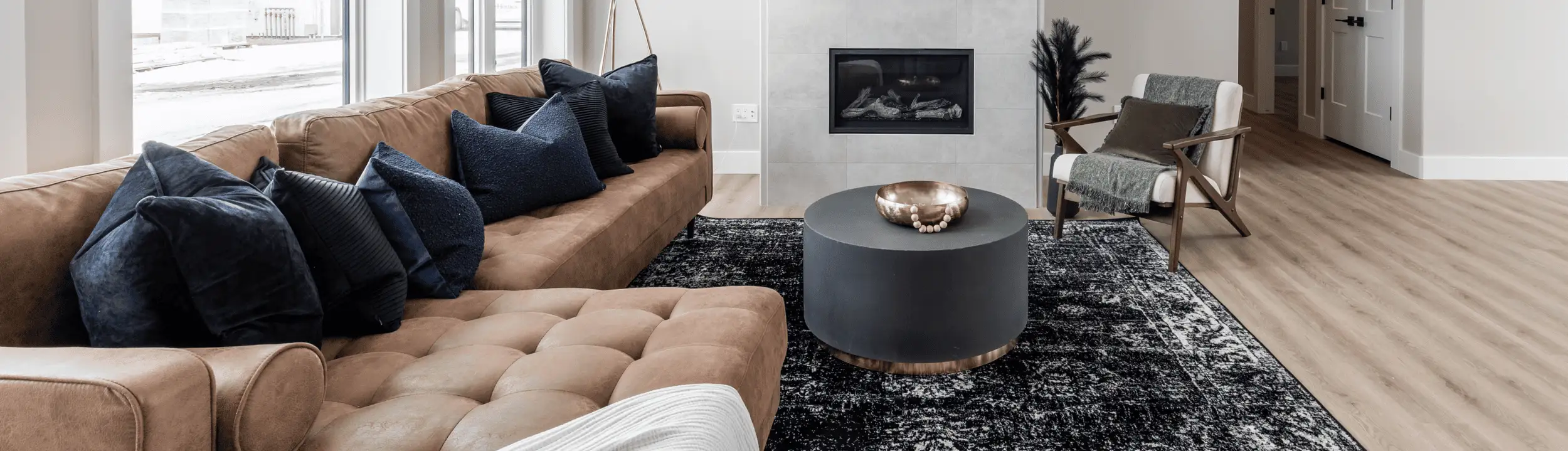 Living room with tan tufted sectional and navy pillows, round charcoal drum coffee table, modern gas fireplace, wood accent chair, and light wood floors on a patterned rug.