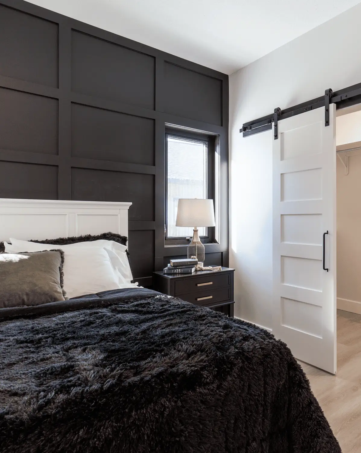 RTM home bedroom in Alberta or Saskatchewan with a dark grid accent wall, white barn door to the closet, white headboard, black plush bedding, and a bedside table with lamp.