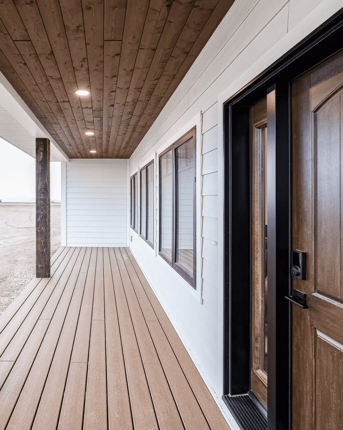 Covered front porch of an RTM home in Alberta or Saskatchewan with wood ceiling, composite decking, white siding, black-trimmed windows, and a wood front door.