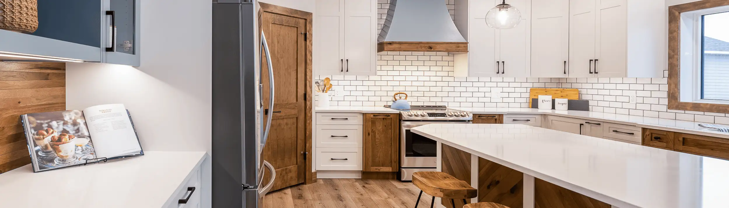 Bright RTM kitchen with white shaker cabinets, subway tile backsplash, quartz island with wood accents and bar stools, custom hood with wood trim, stainless appliances, and a walk-in pantry door.