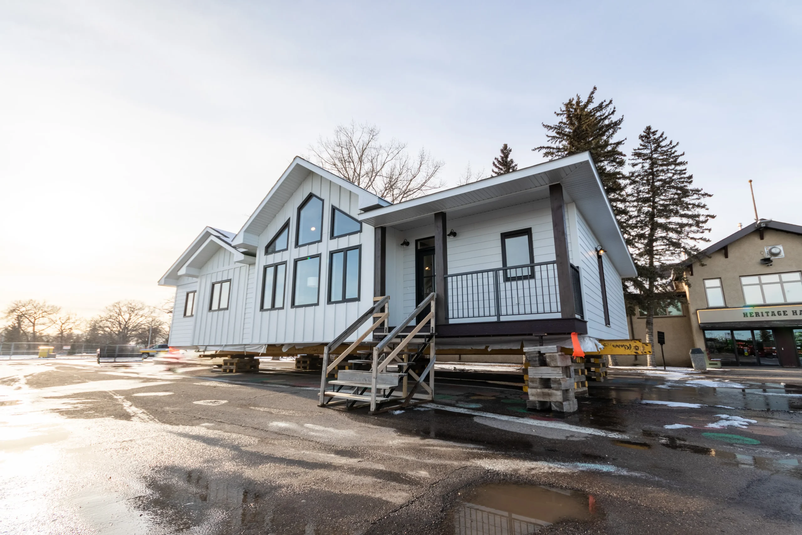 Ready-to-move home elevated on cribbing with white board-and-batten siding, black-framed gable windows, and a covered porch with railing and temporary stairs, staged for transport.