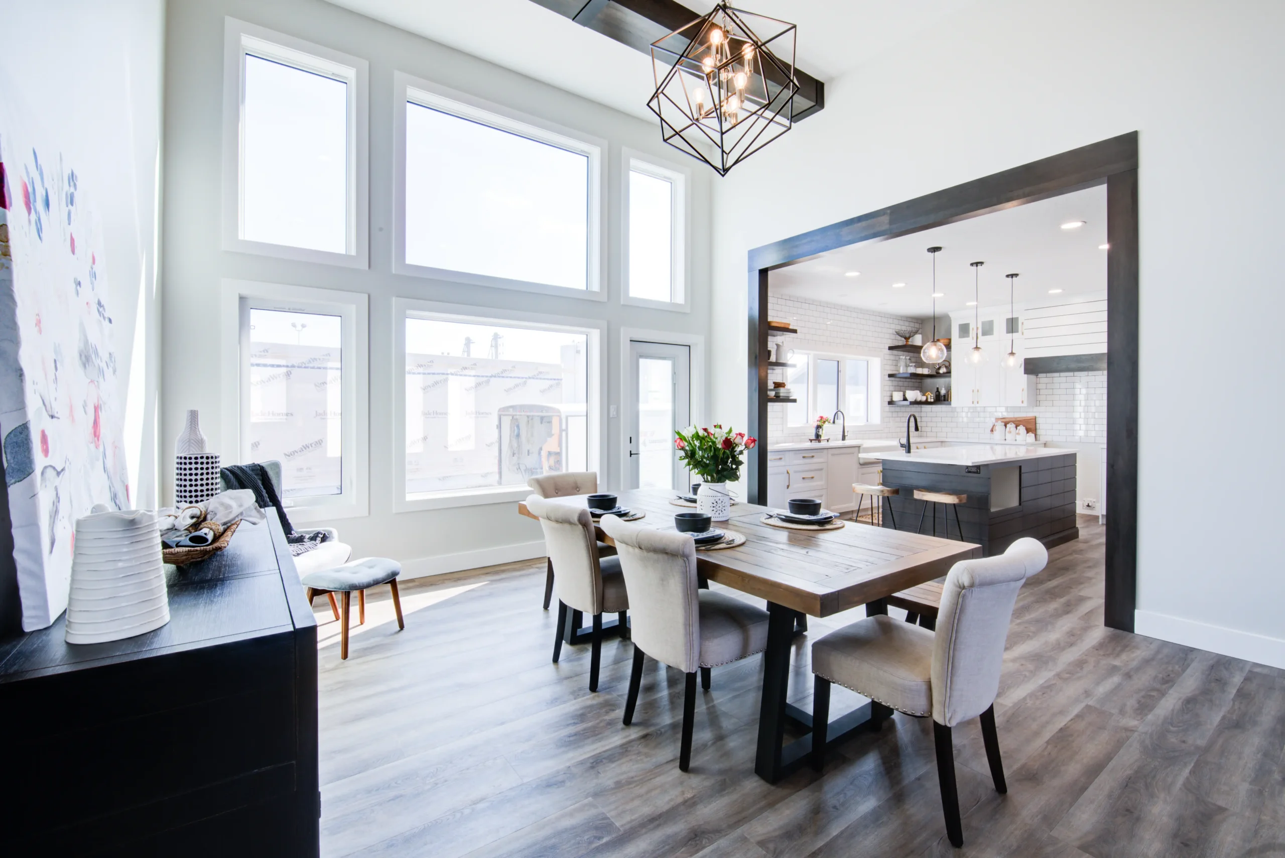 Bright RTM home dining room with large floor-to-ceiling windows, wood dining table with upholstered chairs, and open concept layout flowing into a modern white kitchen with island seating.