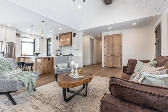 Open-concept living room and kitchen with vaulted shiplap ceiling, white cabinetry, wood range hood and island, brass pendants, wide-plank floors, gray lounge chairs and brown sofa around a live-edge coffee table, and natural wood double doors.