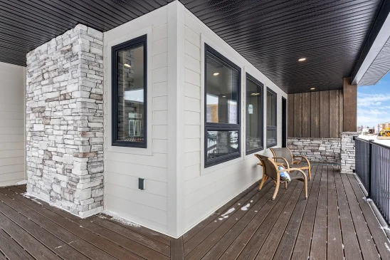 Covered porch of a ready-to-move (RTM) home featuring stone pillars, wood siding, dark trim windows, and composite decking with seating area.