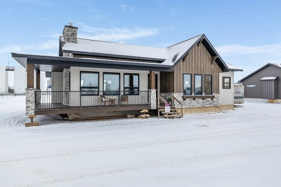Ready-to-move (RTM) home with modern farmhouse design, featuring stone accents, vertical wood siding, large front windows, and a covered porch with black railing, shown in a winter setting.