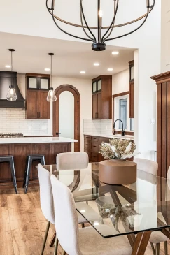 Modern dining area with a glass table and upholstered chairs, overlooking a kitchen with rich wood cabinetry, quartz countertops, and pendant lighting.