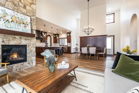 Open-concept living room with stone fireplace, wood coffee table, and modern kitchen and dining area featuring wood cabinetry and pendant lighting.