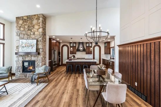 Open-concept living and dining area with stone fireplace, wood accents, and modern kitchen featuring island seating and pendant lighting.