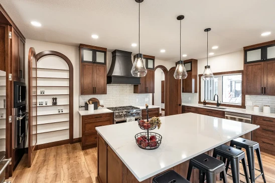 Spacious kitchen with large island, pendant lighting, dark wood cabinets, stainless steel appliances, open arched pantry door with shelving, and subway tile backsplash.