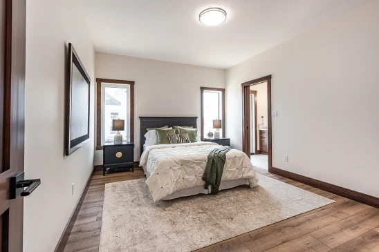 Bright bedroom with wood trim windows, dark nightstands, modern lamps, a bed with white bedding and green accent pillows, and an ensuite bathroom entrance.