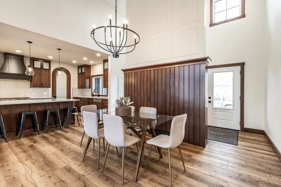 Dining area in an RTM home featuring a glass table with upholstered chairs, wood accent wall, and open kitchen with island and pendant lights.