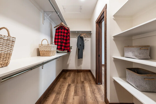 Walk-in closet with wood flooring, white shelves and hanging rods, woven storage baskets, and a red plaid shirt and dark outfit hanging on wooden hangers.