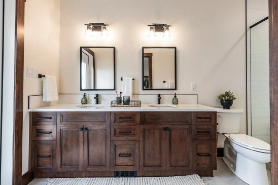 Modern bathroom with double vanity featuring dark wood cabinets, white countertops, two rectangular mirrors, wall-mounted lights, and a toilet with a small plant on top.