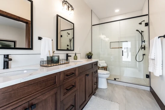 Contemporary bathroom with double vanity, wood cabinets, white countertops, black framed mirrors, glass-enclosed walk-in shower with black fixtures, and a toilet beside a small plant.