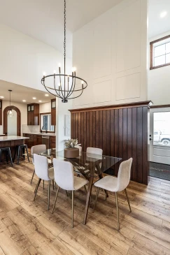Dining area in an RTM home featuring a glass table with upholstered chairs, wood accent wall, and open kitchen with island and pendant lights.