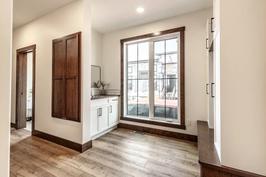 Bright hallway with large window, wood trim, and light wood flooring, featuring white built-in cabinetry with dark hardware and a small vanity area with mirror and plants.