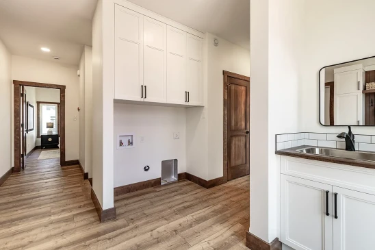 Spacious laundry room with wood flooring, white cabinets, washer and dryer hookups, and a utility sink with a mirror and tiled backsplash.