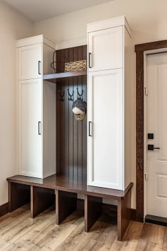 Built-in mudroom storage with white cabinets, dark wood bench and back paneling, cubbies underneath, coat hooks, and a basket and hat on display.