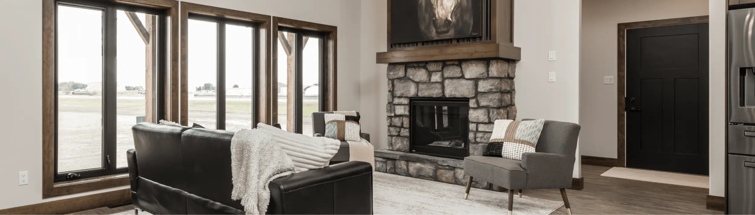 RTM home living room in Alberta or Saskatchewan with large wood-trimmed windows, stone fireplace with horse artwork, black leather sofa, and gray accent chairs.