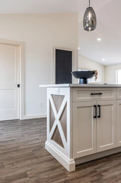 Bright kitchen interior featuring a white island with wood accents, black hardware, and pendant lighting above, set against light walls and wood-grain flooring in a Saskatchewan ready-to-move home.