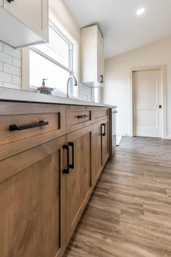 Modern kitchen view showcasing wood lower cabinets with black handles, white quartz countertops, subway tile backsplash, and a large window above the sink in a Saskatchewan ready-to-move home.