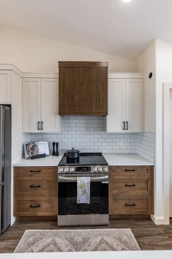 Modern kitchen with stainless steel oven and range, white upper cabinets, wood lower drawers, subway tile backsplash, and a matching wood range hood in a Saskatchewan ready-to-move home.