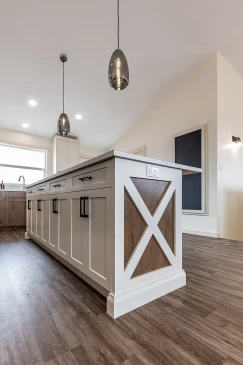 Close-up of a modern farmhouse-style kitchen island with white paneling, wood accents in an X design, quartz countertop, and black hardware, illuminated by pendant lights in a Saskatchewan ready-to-move home.