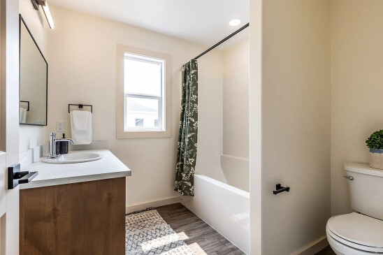 Modern bathroom with light walls, wood vanity, large mirror, white bathtub with green floral shower curtain, and toilet with potted plant accent, in a Saskatchewan ready-to-move home.