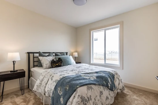 Bright bedroom in a Saskatchewan ready-to-move home featuring a metal bed frame, floral bedding with blue and gray accents, matching side tables with lamps, and a large window letting in natural light.