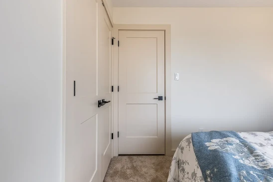Cozy bedroom corner featuring neutral beige doors with modern black hardware, soft carpet flooring, and a bed partially visible with floral bedding and a blue throw blanket.