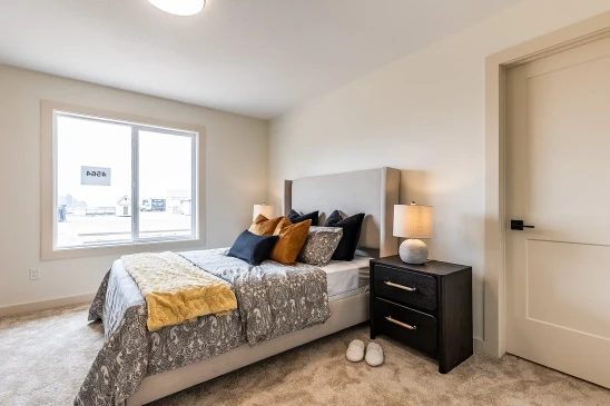 Cozy bedroom in a ready-to-move home featuring a gray upholstered bed with patterned bedding, accent pillows, a dark wood nightstand with a lamp, and a large window letting in natural light.