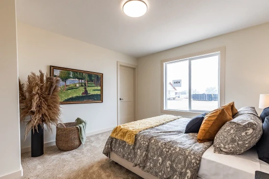 Stylish bedroom in a Saskatchewan ready-to-move home featuring a large window, patterned bedding with warm-toned pillows, framed landscape art, a woven basket with a throw blanket, and tall pampas grass decor.