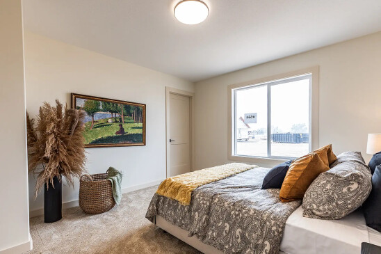 Stylish bedroom in a Saskatchewan ready-to-move home featuring a large window, patterned bedding with warm-toned pillows, framed landscape art, a woven basket with a throw blanket, and tall pampas grass decor.