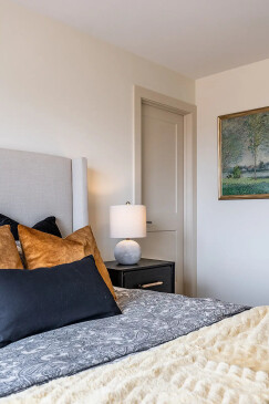 Close-up view of a cozy bedroom featuring a modern upholstered headboard, layered throw pillows in earth tones, a patterned comforter, and a bedside table with a lamp and framed wall art.
