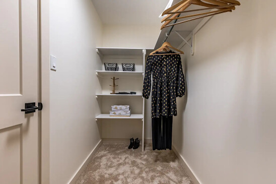 Modern walk-in closet in a ready-to-move home featuring beige walls, carpet flooring, built-in white shelves with baskets and boxes, and a clothing rod with hangers and a black patterned dress.