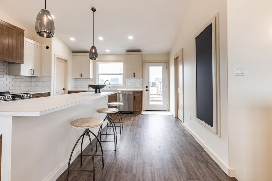 Modern kitchen in a ready-to-move home featuring a long white breakfast bar with wooden stools, two pendant lights, white cabinets with wood accents, and stainless steel appliances.