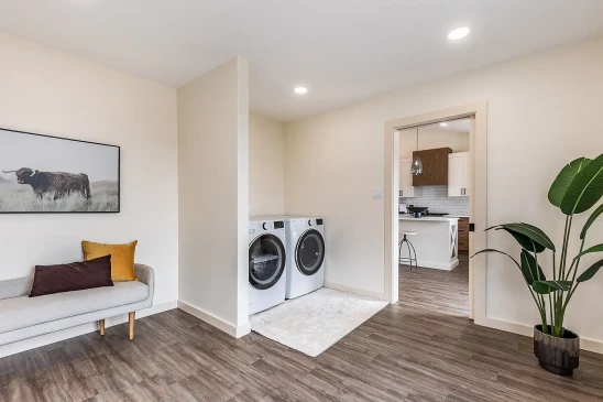 Bright and clean laundry area in a ready-to-move home with modern front-load washer and dryer, a minimalist bench with colorful pillows, and wood-look vinyl flooring.