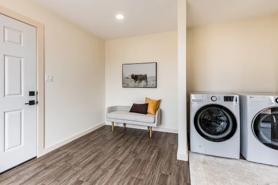 Bright and clean laundry area in a ready-to-move home with modern front-load washer and dryer, a minimalist bench with colorful pillows, and wood-look vinyl flooring.