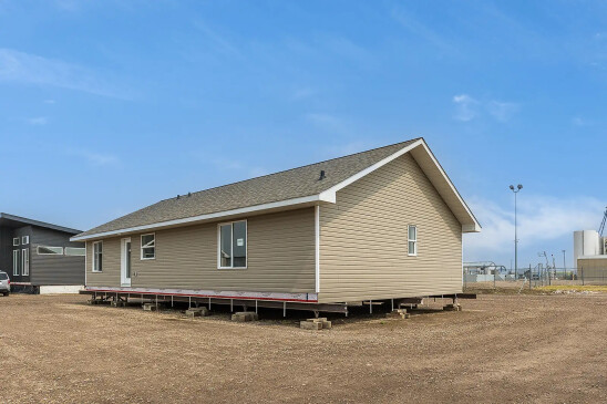 Side view of a tan ready-to-move home in Saskatchewan, elevated on support blocks with white trim, asphalt shingles, and a clear blue sky overhead.