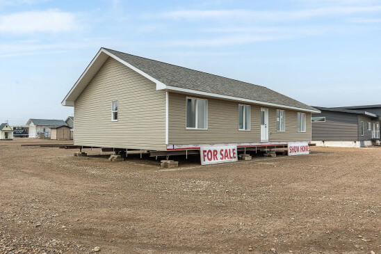 Front view of a tan ready-to-move show home for sale in Saskatchewan, featuring white-trimmed windows, a gable roof, and signs that read “FOR SALE” and “SHOW HOME.”