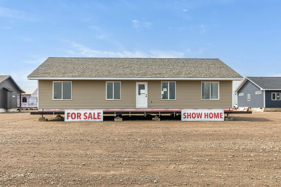 Front view of a tan ready-to-move show home for sale in Saskatchewan, featuring white-trimmed windows, a gable roof, and signs that read “FOR SALE” and “SHOW HOME.”
