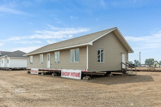 Tan ready-to-move (RTM) show home for sale in Saskatchewan, displayed on a gravel lot with white-trimmed windows and a gable roof under a bright blue sky.